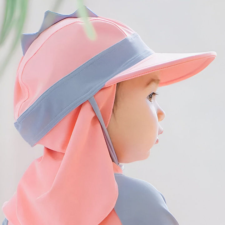Deux adorables bébés en maillots de bain anti-UV colorés avec chapeaux assortis. L'un en rose et gris, l'autre en turquoise et gris, posant devant un mur blanc et un plancher en bois clair. Vêtements de plage pour enfants, protection solaire pour bébés.