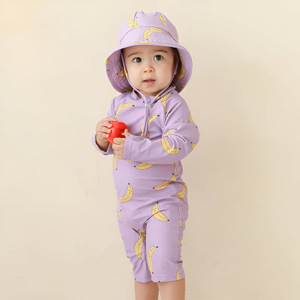 Bébé portant un maillot de bain et un chapeau lilas imprimés de bananes, tenant un jouet rouge. Vêtement de plage pour enfant, protection solaire.