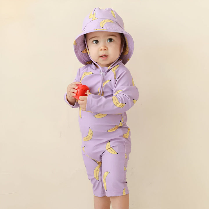 Bébé portant un maillot de bain et un chapeau lilas imprimés de bananes, tenant un jouet rouge. Vêtement de plage pour enfant, protection solaire.