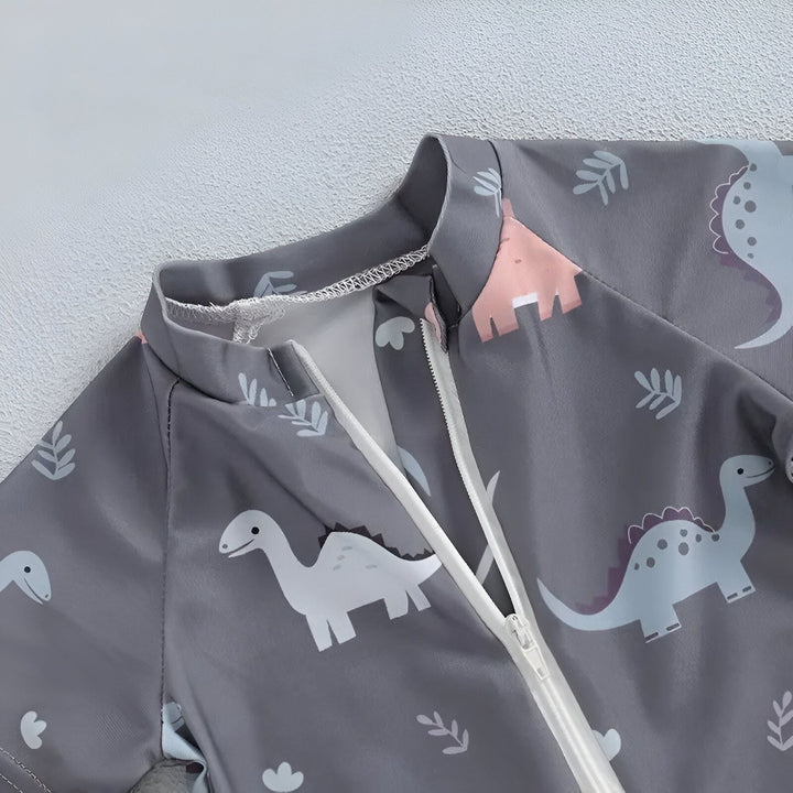 Gros plan sur un vêtement de bébé gris avec des motifs de dinosaures de différentes couleurs et des motifs de feuilles. On peut apercevoir une fermeture éclair blanche.