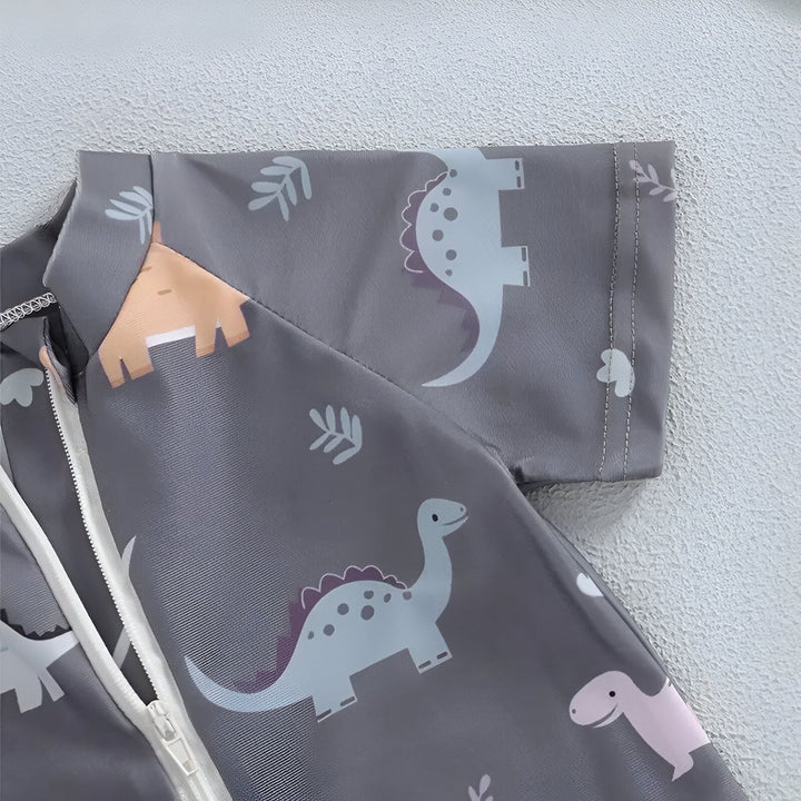 Gros plan d'un maillot de bain gris à manches longues pour enfant, orné de motifs de dinosaures et de feuilles. Fermeture éclair blanche visible. Idéal pour protéger les enfants du soleil avec style.
