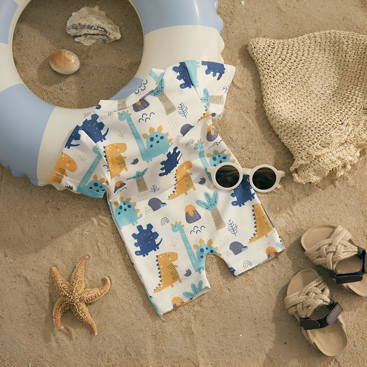 Gros plan d'un vêtement pour enfant à motifs de dinosaures bleus et jaunes sur un fond blanc. Le vêtement est posé sur du sable, créant une ambiance ludique et estivale. Idéal pour les petits explorateurs en herbe.