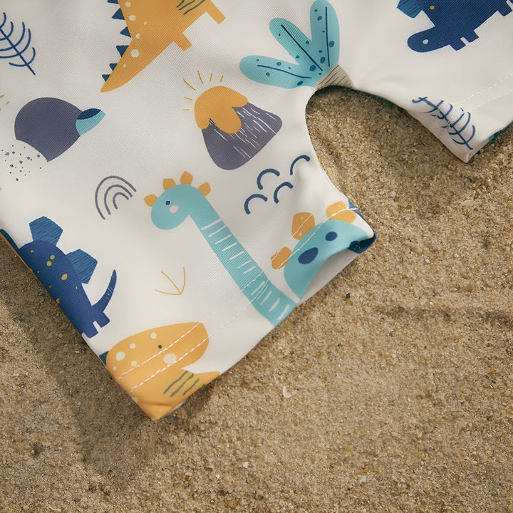 Gros plan sur un tissu blanc imprimé de motifs enfantins : dinosaures bleus et orange, volcans jaunes et plantes turquoise. Texture douce et colorée, idéale pour une chambre d'enfant.