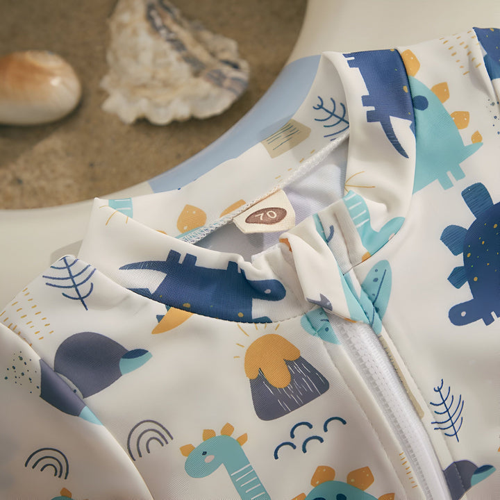 Gros plan sur un maillot de bain bébé à manches longues, imprimé de dinosaures et de motifs amusants. Fermeture éclair blanche visible. Coquillages décoratifs à l'arrière-plan.