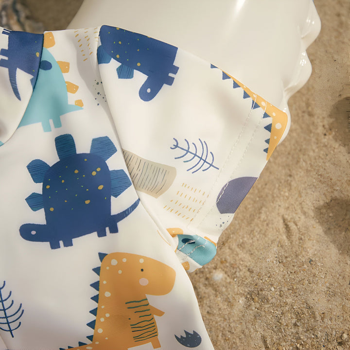 Ensemble de plage pour enfants avec motif dinosaure : maillot de bain, bouée, chapeau de paille, lunettes de soleil et sandales sur le sable. Ambiance estivale et vacances à la mer pour les petits.