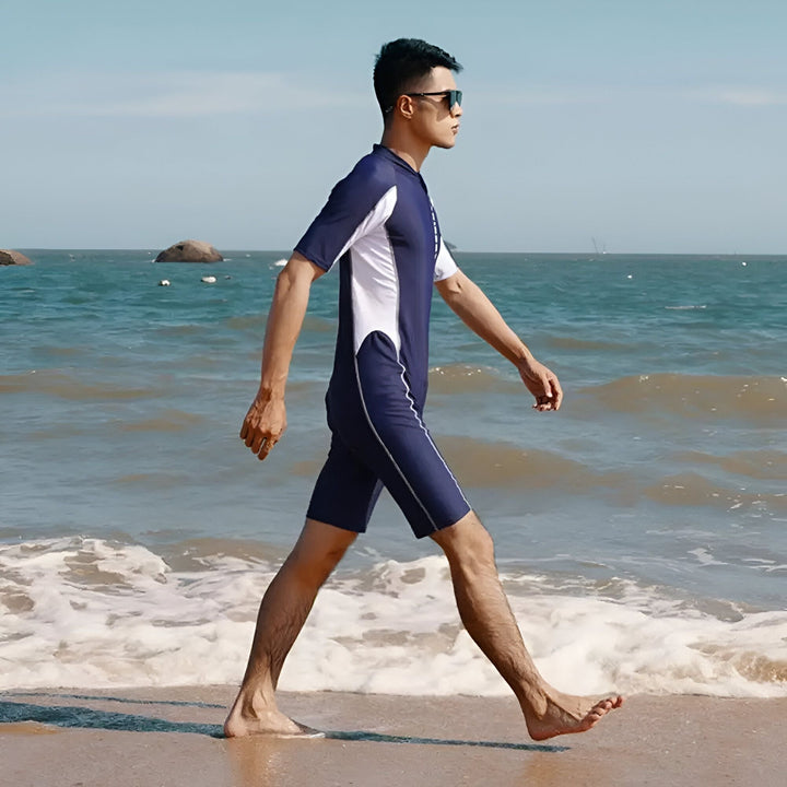 Homme portant une combinaison de bain bleu marine et blanche à manches courtes, lunettes de soleil noires, debout dans l'eau à la plage. Mer et ciel bleu en arrière-plan.