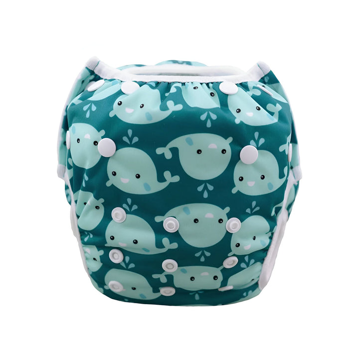 Bébé en couche de bain, motif baleines bleues, près d'une piscine. Il se tient sur la pointe des pieds, explorant le bord. Lumière naturelle et ambiance estivale.