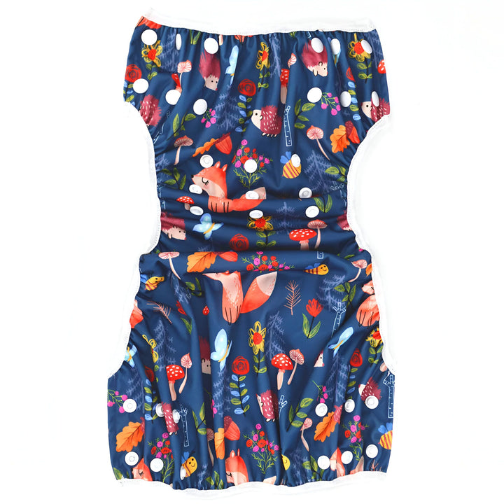 Couche lavable avec un motif de forêt enchantée. On voit des renards, des champignons, des hérissons et des feuilles sur un fond bleu marine. Des boutons pressions blancs servent de fermeture.