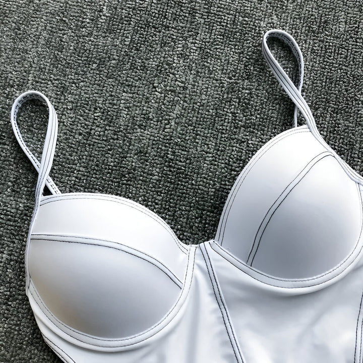 Image d'un maillot de bain une pièce moderne et bicolore (blanc et noir) avec des bretelles fines, posé sur une surface texturée grise. À côté, on aperçoit un petit sac jaune et une petite branche décorative blanche, ajoutant une touche esthétique.