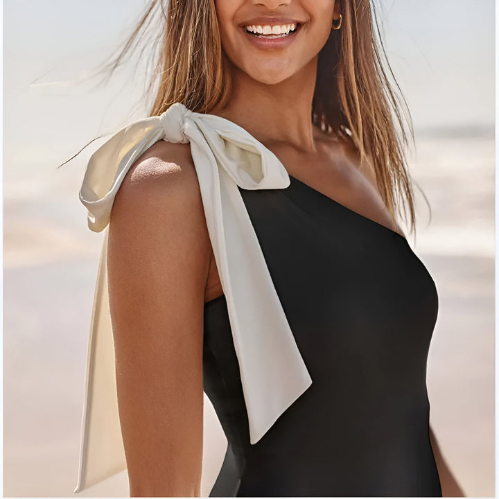 Femme souriante portant un maillot de bain une pièce noir avec une épaule dénudée et un nœud blanc sur l'épaule. Mer en arrière-plan. Mode de plage, été.