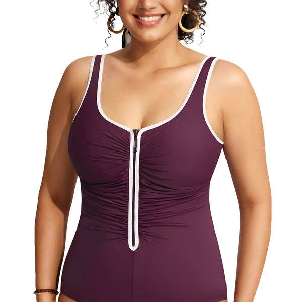 Image d'une femme souriante portant un maillot de bain une pièce prune avec une fermeture éclair blanche sur le devant et un liseré blanc autour du cou et des bras. Elle porte des boucles d'oreilles dorées et un bracelet.