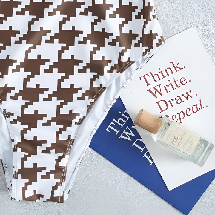 Image d'un maillot de bain une pièce à motif pied-de-poule marron et blanc, avec chapeau de paille et lunettes de soleil blanches.