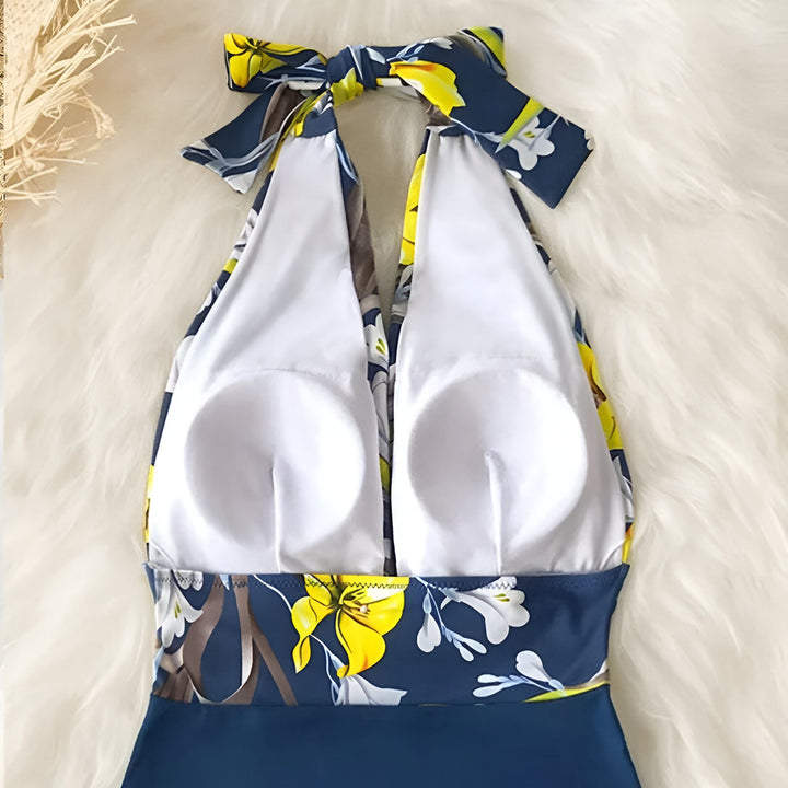 Vue arrière d'une femme à la plage portant un maillot de bain une pièce bleu et motif floral, avec un chapeau de paille et un foulard dans les cheveux. Mer et rochers en arrière-plan.