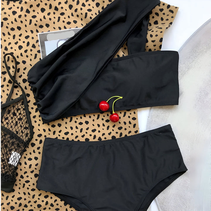 Femme portant un maillot de bain noir une pièce à une épaule avec un bracelet doré au poignet, posant devant un rideau blanc.