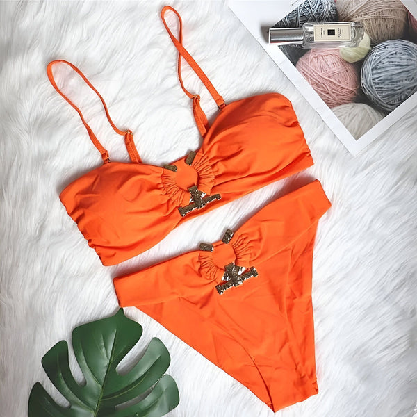 Image d'un ensemble de bikini orange vif avec des détails dorés, présenté à plat sur un fond texturé blanc. On voit également un parfum et des pelotes de laine aux couleurs pastel en arrière-plan.