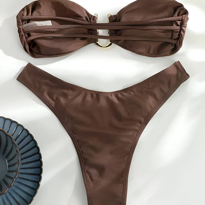 Femme en bikini marron avec bijoux dorés sur une plage de sable blanc. Maillot de bain deux pièces, chaîne dorée autour du cou, bracelets aux poignets et bagues aux doigts. Mode estivale, style de plage.