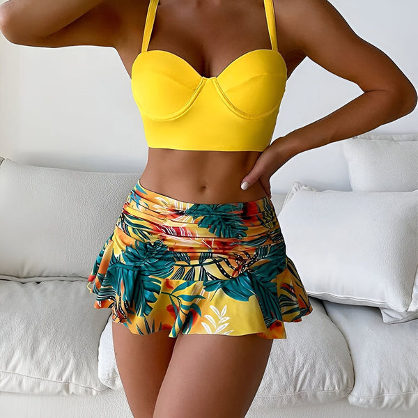 Une femme vue de dos, portant un haut de bikini orange et une jupe courte à motifs floraux rouges et bleus. Elle pose devant un mur blanc. Mode estivale, vêtements de plage.