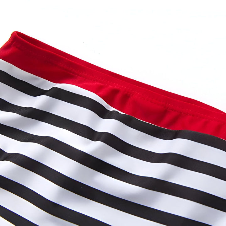 Maillot de bain deux pièces pour enfant. Haut triangle blanc à bordure rouge avec liens rouges à nouer dans le cou. Bas rayé noir et blanc, ceinture rouge. Étoile de mer et coquillage à côté.