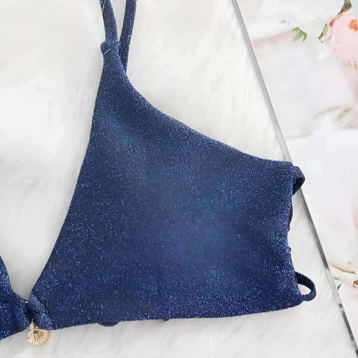 Haut de bikini bleu scintillant avec fines bretelles sur une surface duveteuse blanche. Détail coquillage doré.