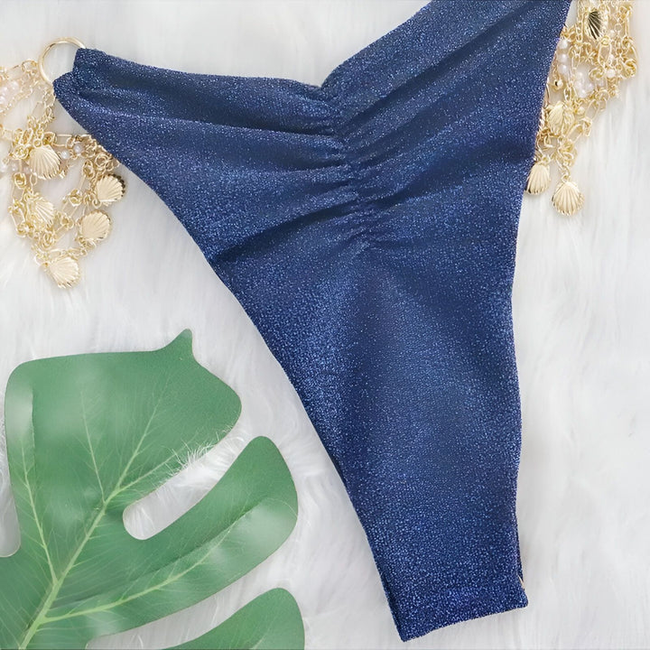 Photo d'un bikini deux pièces bleu marine brillant avec ornements dorés, posé sur une fourrure blanche douce. Haut triangle à bretelles fines, bas échancré avec chaînes décoratives. Élégance estivale.
