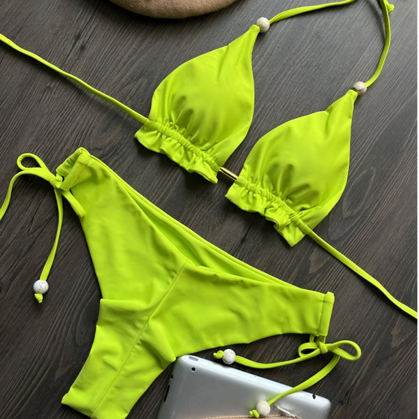 Vue rapprochée d'un bikini vert olive à fines bretelles et d'un béret beige à perles, posés sur un fond en bois foncé. Style élégant pour l'été.