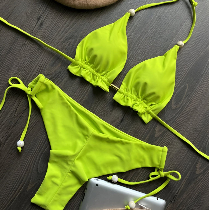 Vue rapprochée d'un bikini vert olive à fines bretelles et d'un béret beige à perles, posés sur un fond en bois foncé. Style élégant pour l'été.