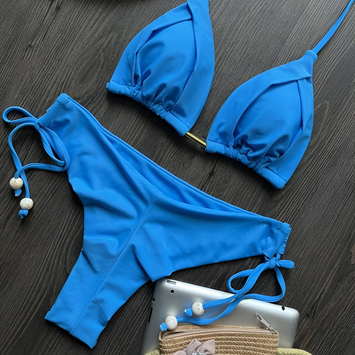 Vue rapprochée d'un bikini bleu turquoise avec un haut triangle et une culotte à nouer sur une surface en bois sombre, avec une tablette et un sac de plage en osier à proximité. Mode estivale, vacances à la plage.