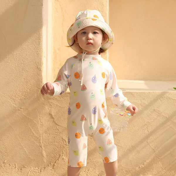 Enfant portant un maillot de bain une pièce à motifs de fruits et un chapeau assorti, debout devant un mur beige. Vêtements de plage pour bébé, protection solaire, mode enfantine.