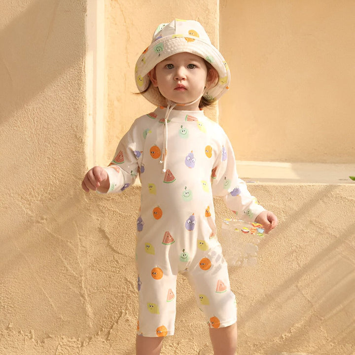 Enfant portant un maillot de bain une pièce à motifs de fruits et un chapeau assorti, debout devant un mur beige. Vêtements de plage pour bébé, protection solaire, mode enfantine.