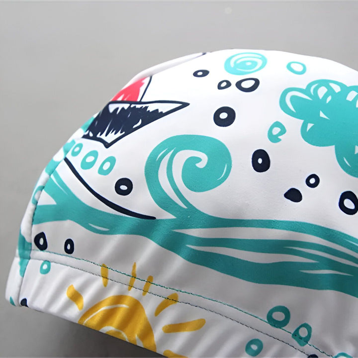 Ensemble de bain pour enfant comprenant un haut à manches courtes imprimé mer, un slip de bain turquoise, un bonnet de bain assorti et des lunettes de soleil bleues. Un coussin en forme de cygne "Echo Park" complète la scène.