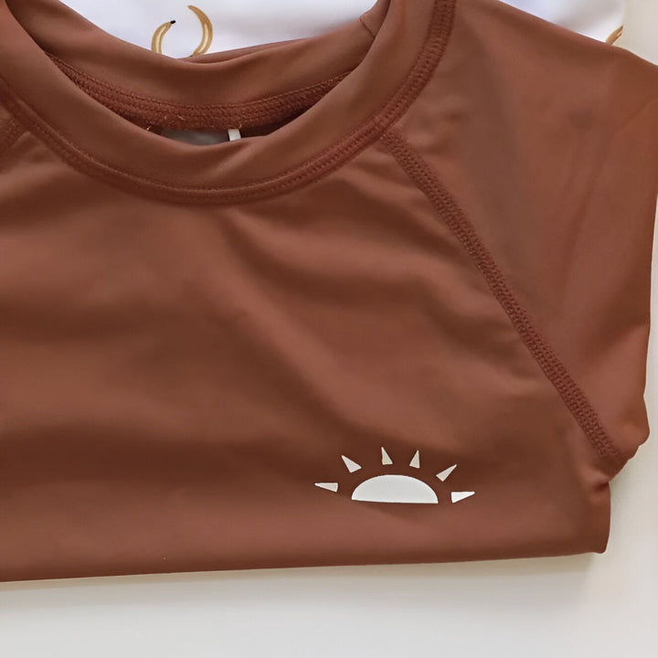 Ensemble de bain pour enfant comprenant un haut à manches longues marron avec un motif de soleil, un short de bain à pois marron, un bonnet de bain assorti et des lunettes de soleil noires. Vêtements d'été pour enfants.