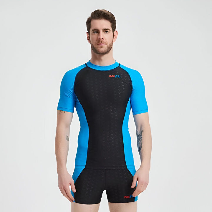 Un homme portant un maillot de bain à manches courtes noir et bleu. Le maillot est moulant et comprend un short de bain assorti. Le fond est blanc et uni.