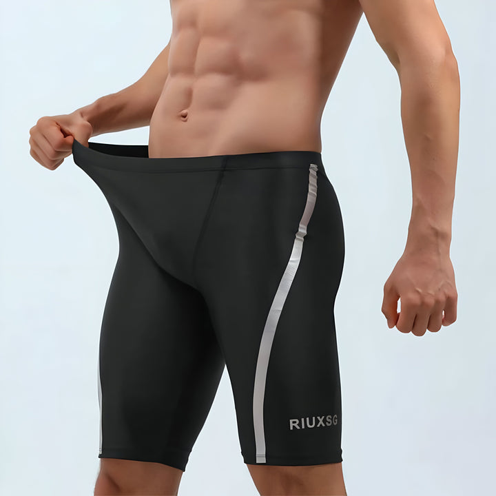 Homme en tenue de sport noire et grise, montrant l'élasticité du tissu du t-shirt. Vêtement de sport moulant avec logo ARXSGI. Short noir avec bande latérale. Idéal pour entraînement, fitness et activités sportives.