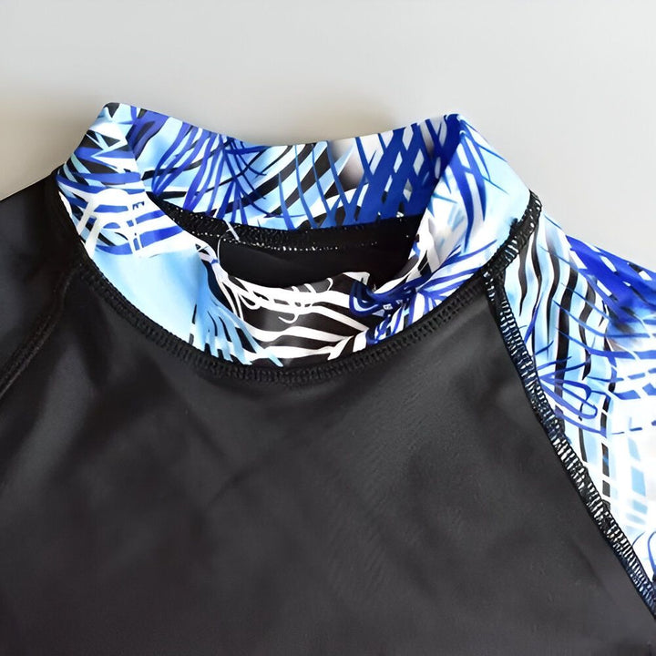 Image d'un rashguard noir à manches longues, avec un imprimé bleu et blanc de feuilles de palmier sur une manche et le logo "TUBETREE" sur le devant.