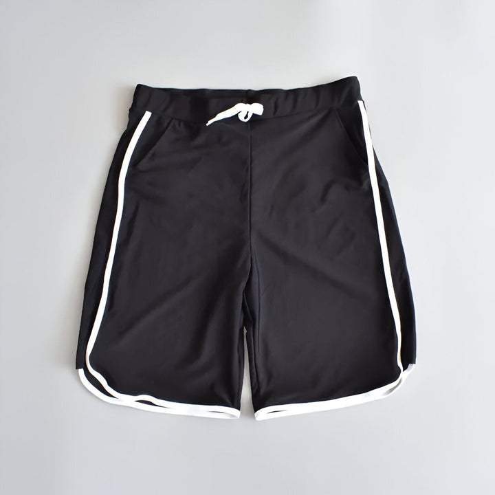 Homme à la plage portant un rashguard Tube Tree noir et à motifs de feuilles de palmier, des lunettes de soleil et un short de bain noir. Arrière-plan : plage et eau turquoise.