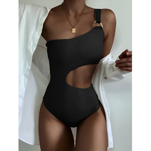 Femme portant un maillot de bain une pièce beige avec une découpe à la taille, une bretelle asymétrique avec un anneau décoratif. Elle porte une chemise blanche ouverte et un collier doré.