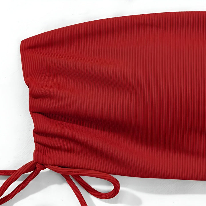 Une femme posant en bikini rose vif. Haut de bikini bandeau sans bretelles, avec une culotte à cordon de serrage. Elle est allongée sur un coussin blanc. L'ambiance est décontractée et estivale.