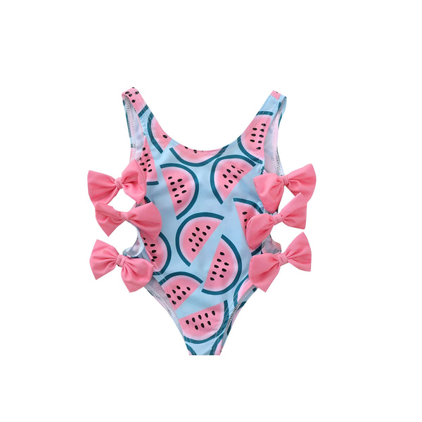 Image d'un maillot de bain une pièce pour enfant, bleu avec des motifs de pastèque et décoré de nœuds roses, accompagné de lunettes de soleil, d'une bouée et de jouets de plage.