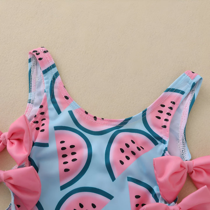Image d'un maillot de bain une pièce pour bébé, bleu avec des motifs de melon d'eau roses, décoré de nœuds roses sur les côtés, avec des lunettes de soleil blanches, une bouée de sauvetage et des jouets sur un fond beige.