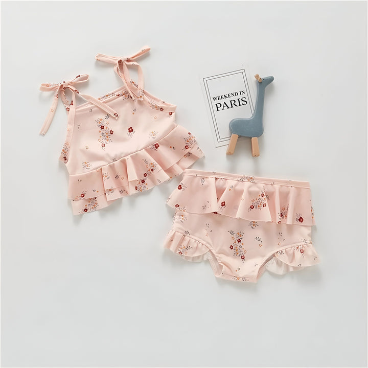 Maillot de bain une pièce rose pâle pour bébé, orné de fleurs, avec volants et bretelles à nouer. Idéal pour les vacances d'été à la plage ou à la piscine.