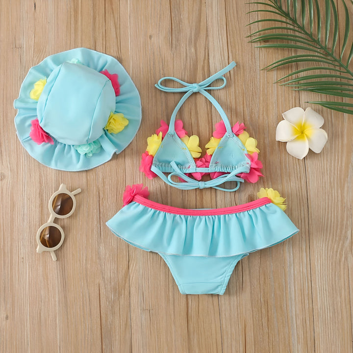 Ensemble de maillot de bain rose vif pour enfant avec un chapeau assorti, orné de fleurs bleues et jaunes. Lunettes de soleil à monture beige complètent le look estival. Feuille de palmier en arrière-plan. Idéal pour les journées à la plage.