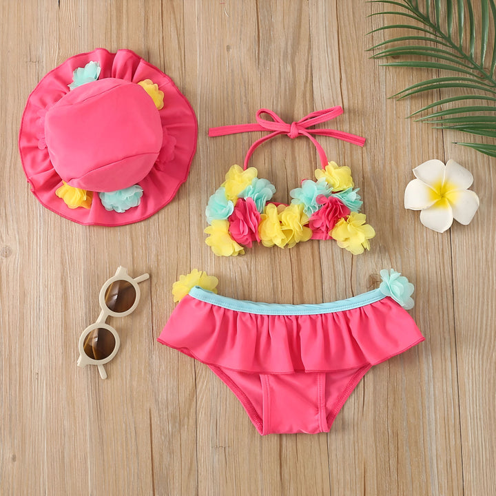 Ensemble de bain pour enfants rose vif avec chapeau, haut de bikini à fleurs colorées et bas à volants, lunettes de soleil rondes et fleur blanche exotique sur fond de bois clair. Vêtements d'été tendance pour filles.