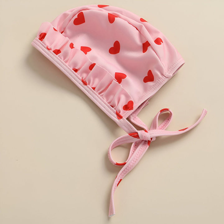 Bonnet de bain rose à motifs de cœurs rouges, avec des volants sur le bord et des liens à nouer. Idéal pour les bébés et les jeunes enfants.