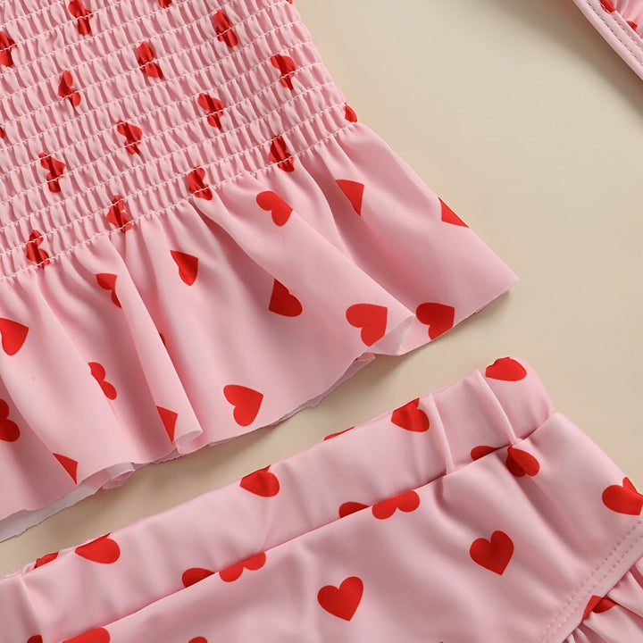 Ensemble de bain rose à motifs de cœurs rouges pour enfant, comprenant un haut smocké à bretelles nouées, une culotte à volants et un bonnet de bain assorti, parfait pour les vacances à la plage et les jeux aquatiques estivaux.