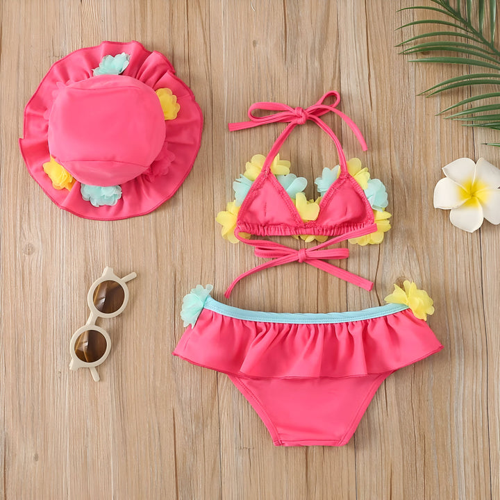 Ensemble de plage pour bébé rose vif avec chapeau, maillot de bain à fleurs et lunettes de soleil rondes sur fond bois. Idéal pour la mode enfantine, les vacances d'été et la protection solaire.