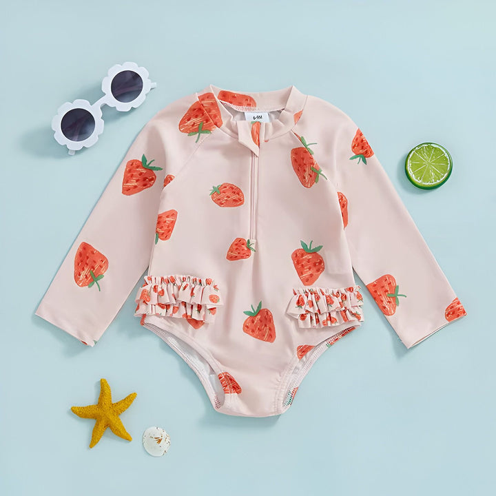 Combinaison de bain bébé rose à manches longues avec motif fraises, fermeture éclair sur le devant, détails à volants, accompagnée de lunettes de soleil blanches en forme de fleurs, étoile de mer jaune et rondelle de citron.