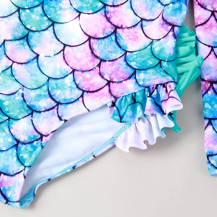 Gros plan d'un maillot de bain une pièce pour enfant avec un motif d'écailles de sirène turquoise et rose, agrémenté de volants. Idéal pour la piscine ou la plage.