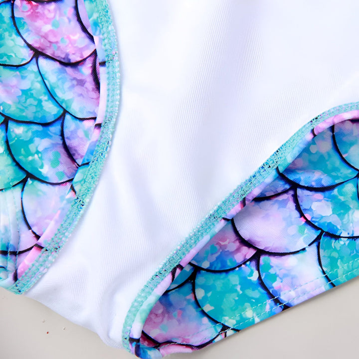 Maillot de bain une pièce à manches longues pour enfant, motif écailles de sirène aux couleurs pastel (bleu, rose, violet). Volants turquoise et blancs. Accessoires et décorations marines.