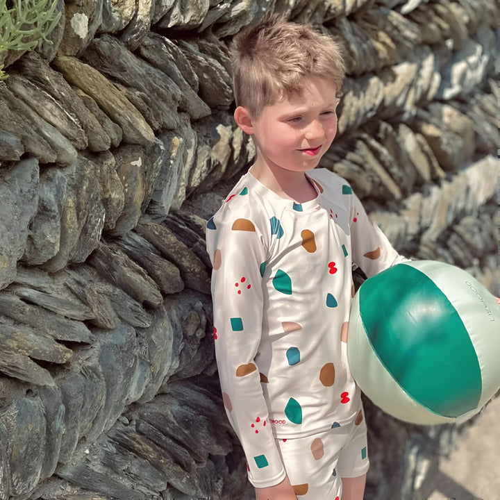 Ensemble de bain pour enfant, comprenant un haut à manches longues et un short assorti. Imprimé géométrique multicolore sur fond beige. Idéal pour la plage et la piscine.