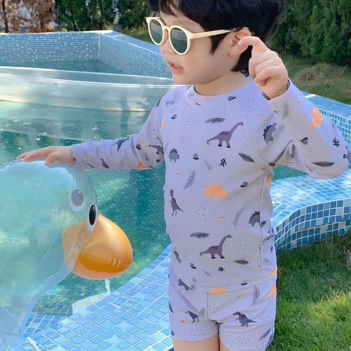 Ensemble de plage pour enfant couleur crème, motif palmiers blancs : haut à manches longues et short assorti. Chapeau de paille et lunettes de soleil rondes complètent le look estival.
